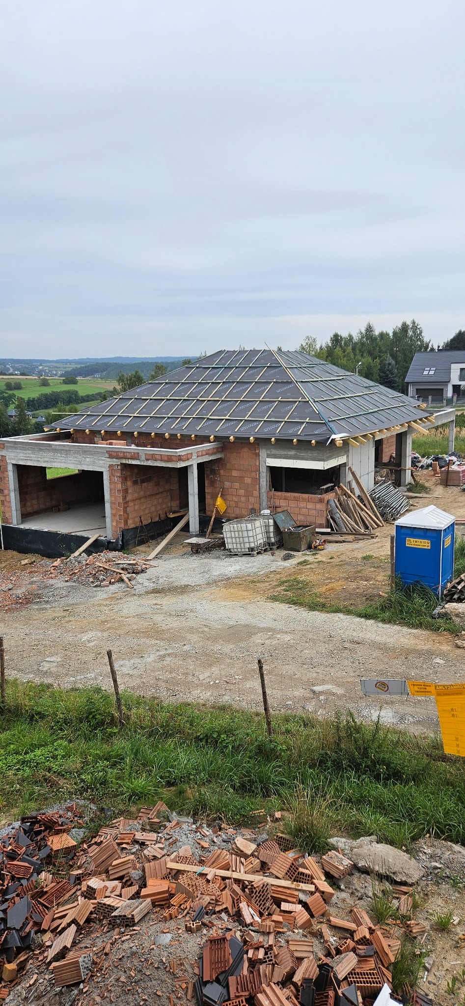 Budowa domu jednorodzinnego w Krakowie, widok z góry na częściowo ukończony budynek z czerwonej cegły i dachem pokrytym membraną. W tle zielony krajobraz.
