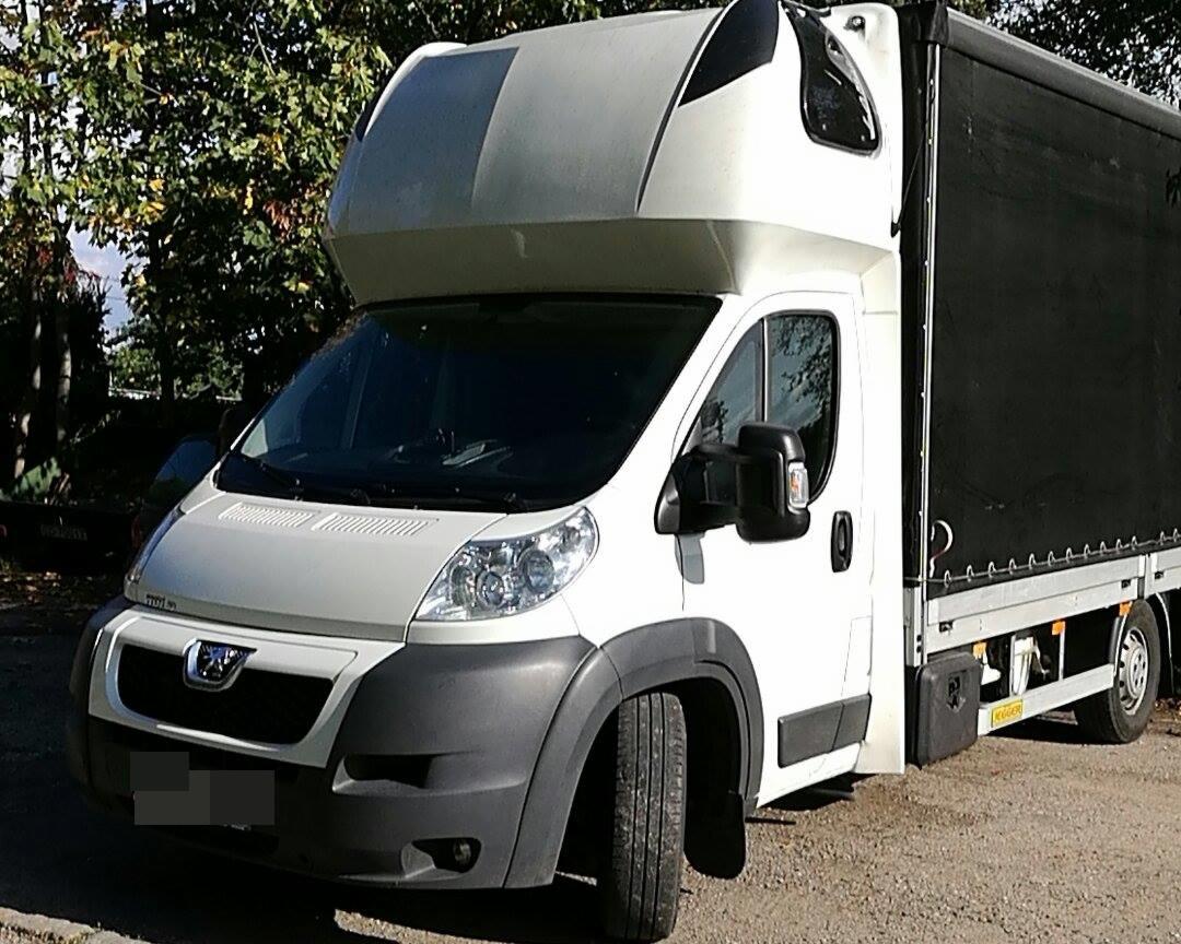 Biały bus dostawczy z czarną plandeką na naczepie, zaparkowany na żwirowym podłożu, z widocznym logo Peugeot na masce.