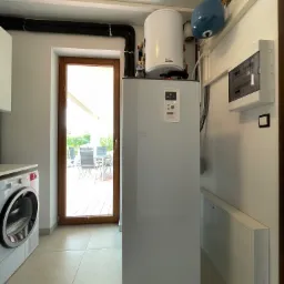 Biała pompa ciepła z zasobnikiem ciepłej wody, zainstalowana w pomieszczeniu gospodarczym obok pralki i drzwi tarasowych.