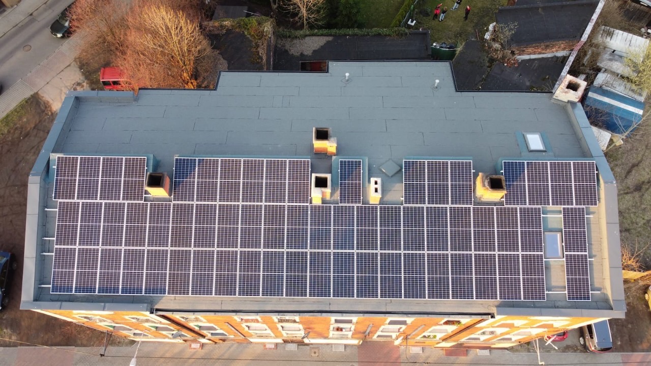 Widok z góry na dach budynku z zainstalowanymi panelami fotowoltaicznymi; widoczne kominy i okna dachowe.