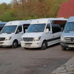 Flota czterech busów Mercedes-Benz Sprinter, dwa srebrne i dwa białe, zaparkowane na żwirowym parkingu przed budynkiem z czerwoną dachówką i zielenią w tle, z otwartymi drzwiami w jednym...