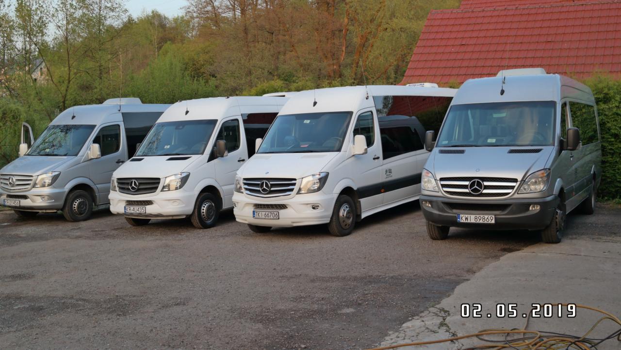 Flota czterech busów Mercedes-Benz Sprinter, dwa srebrne i dwa białe, zaparkowane na żwirowym parkingu przed budynkiem z czerwoną dachówką i zielenią w tle, z otwartymi drzwiami w jednym...