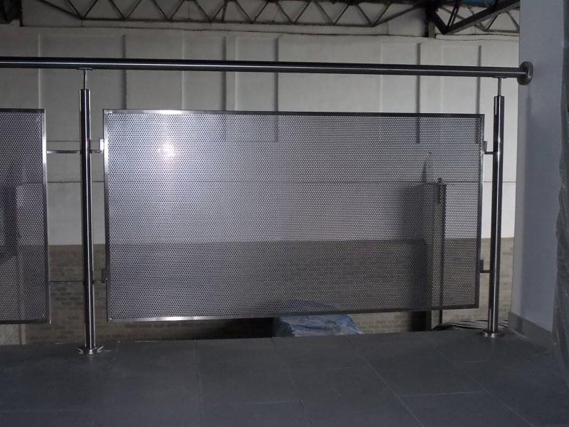 Metalowa balustrada z perforowanej blachy, mocowana na stalowych słupkach, widoczna na tle szarej ściany wewnątrz budynku.