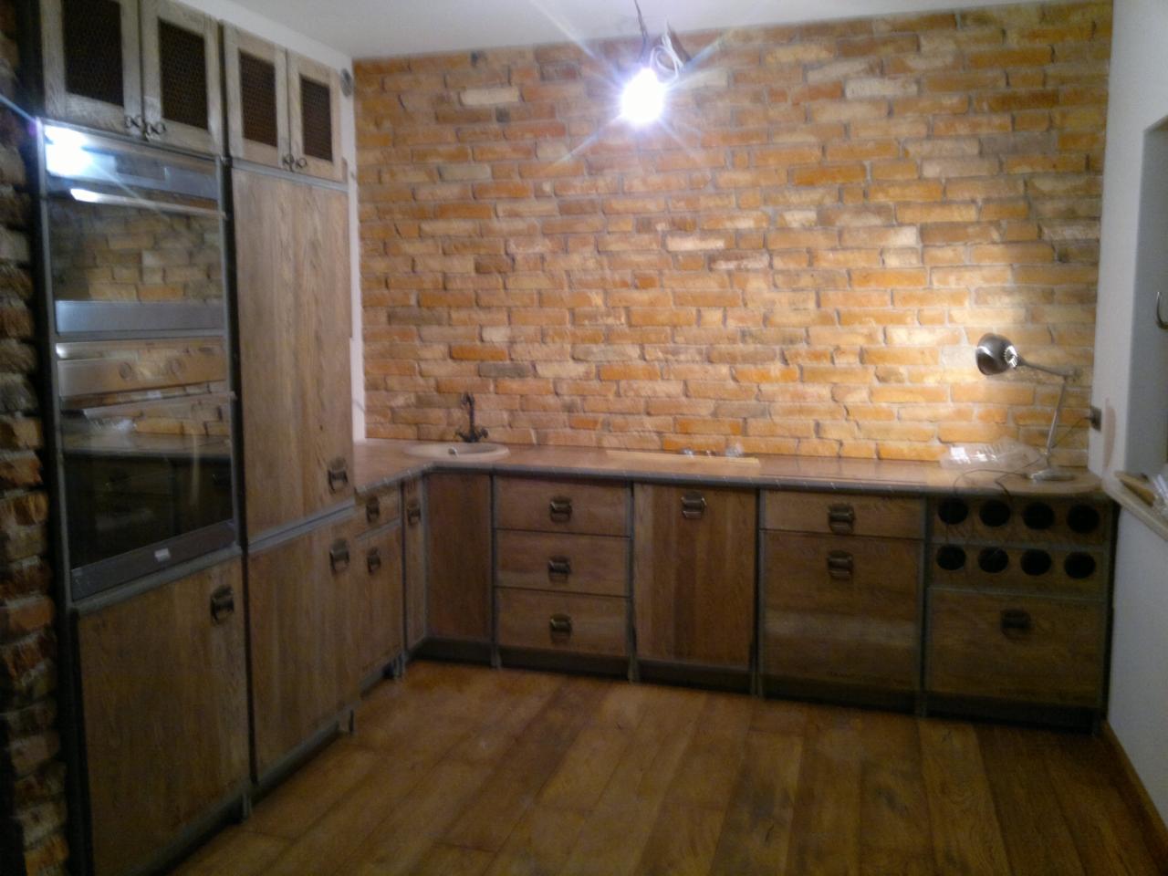 Rustykalna kuchnia z drewnianymi szafkami, ceglaną ścianą i drewnianą podłogą w ciepłym oświetleniu, widoczny fragment blatu z umywalką i metalową lampką.