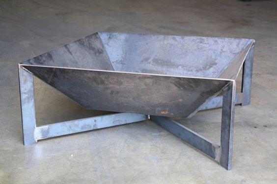 Surowa, kwadratowa misa paleniskowa wykonana z grubej blachy stalowej, wsparta na spawanych nogach z profili, eksponowana na betonowej posadzce.