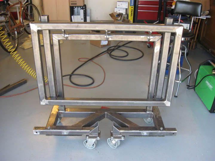 Stalowa konstrukcja ramy na wózku z kółkami, spawana, widoczny sprzęt spawalniczy w tle, garażowe otoczenie.