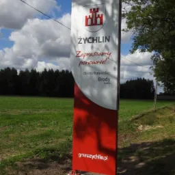 Stojący pionowy szyld informacyjny z herbem i napisem Żychlin, zapraszający ponownie, zamontowany na betonowej podstawie na trawiastym terenie.