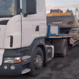 Biała ciężarówka Scania z naczepą przewożąca żółto-czarną koparkę Volvo na tle pochmurnego nieba.