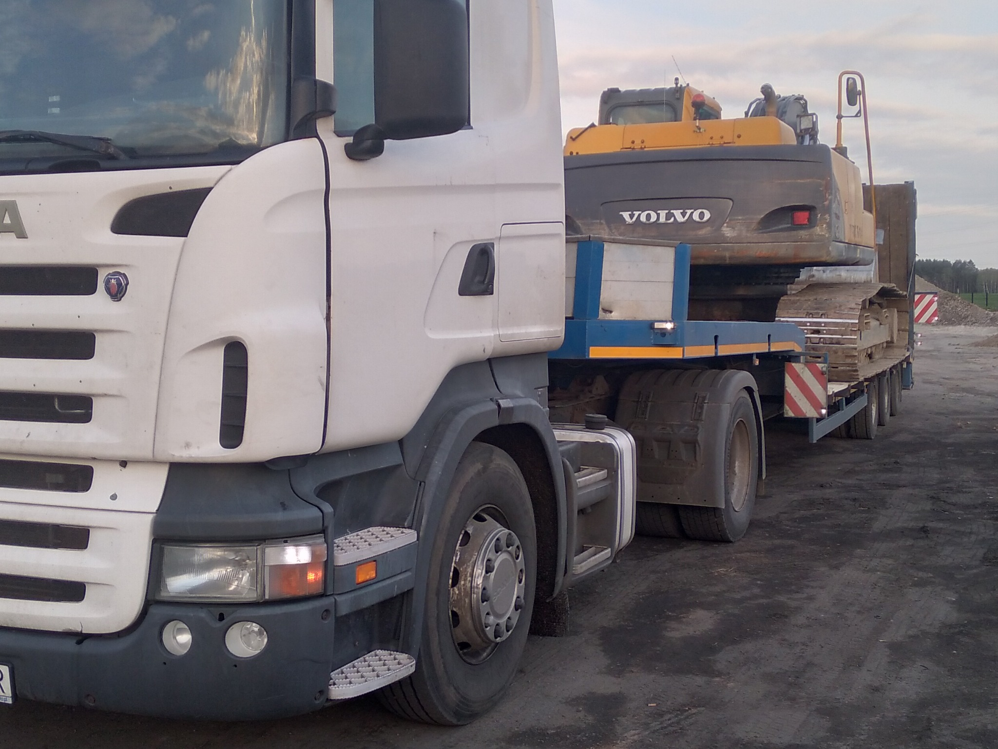 Biała ciężarówka Scania z naczepą przewożąca żółto-czarną koparkę Volvo na tle pochmurnego nieba.