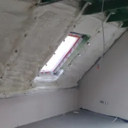 Poddasze z oknem dachowym po izolacji pianką, widoczne nierównomierne pokrycie, fragmenty więźby dachowej i surowy stan wykończenia wnętrza.