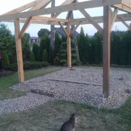 Konstrukcja drewnianej altany w trakcie budowy, podłoże wyłożone kamieniami, w kadrze siedzący kot.