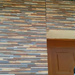 Fragment elewacji budynku z dekoracyjną okładziną z płytek imitujących kamień w odcieniach brązu, beżu i szarości, widoczna drewniana rama drzwi wejściowych z fragmentem ościeżnicy.