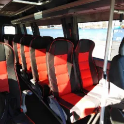 Wnętrze autobusu z czerwono-czarnymi skórzanymi siedzeniami, widok na rzekę przez okna, poręcze wzdłuż przejścia.