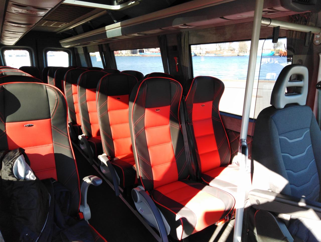 Wnętrze autobusu z czerwono-czarnymi skórzanymi siedzeniami, widok na rzekę przez okna, poręcze wzdłuż przejścia.