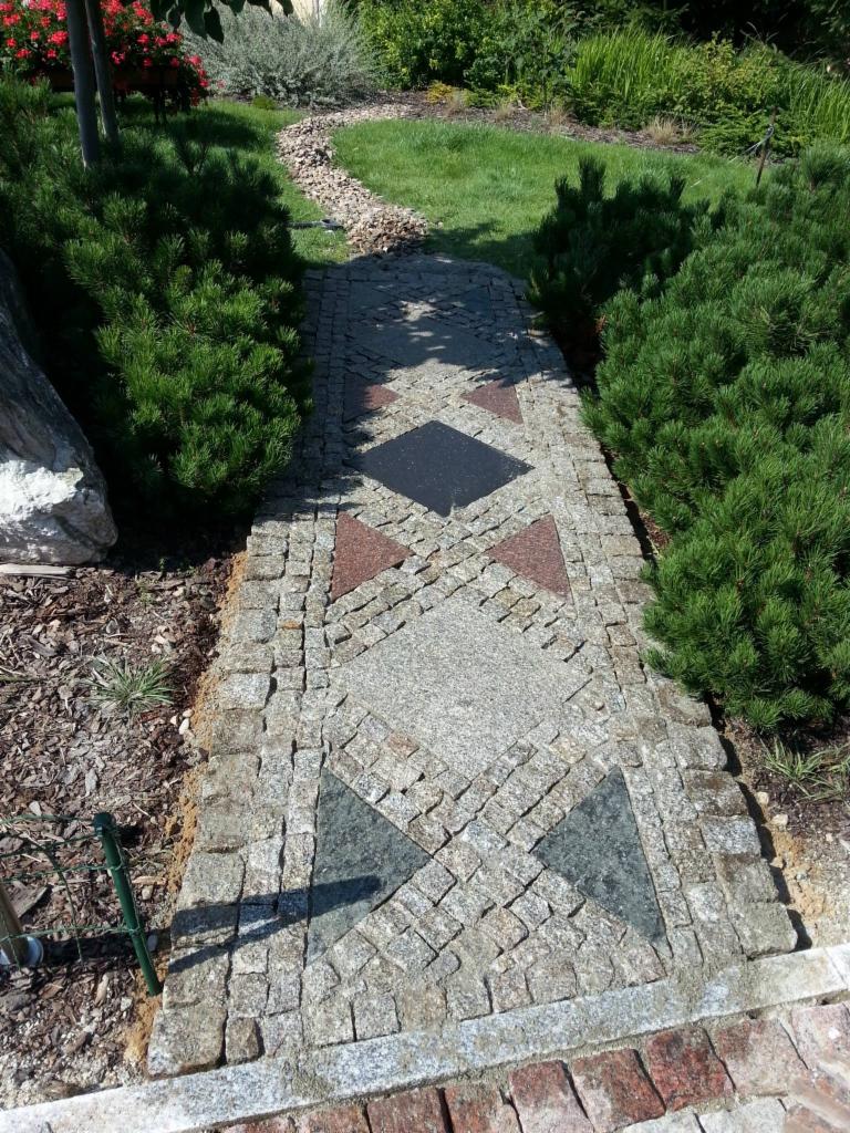 Ścieżka w ogrodzie wyłożona kostką brukową w geometryczny wzór z elementami w kształcie rombów i trójkątów z kamienia w różnych kolorach, otoczona zielenią.