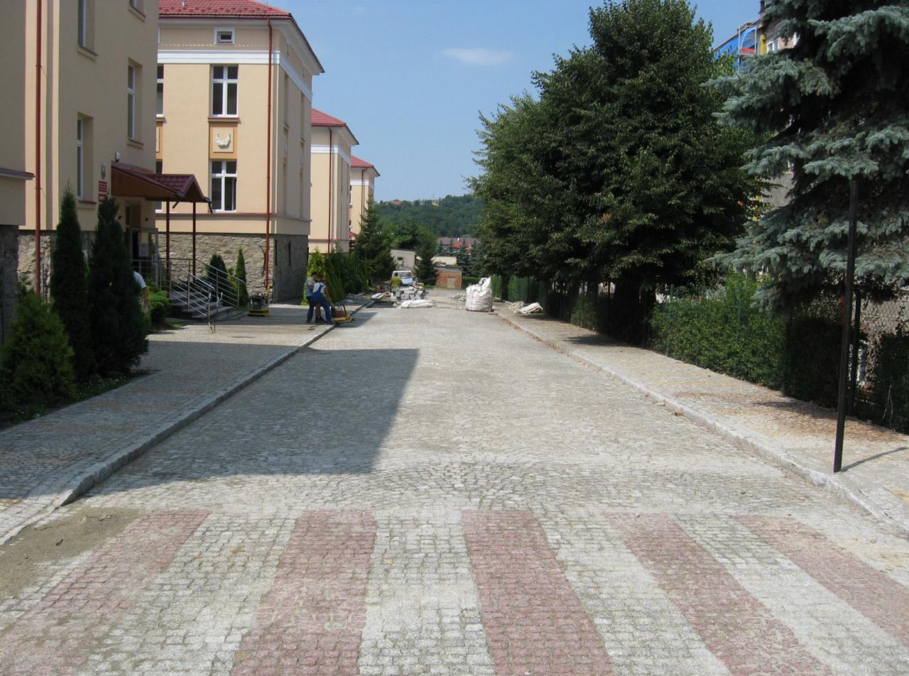 Ulica wyłożona kostką brukową z czerwonymi pasami przejścia dla pieszych, dwóch pracowników w oddali używa zagęszczarki, a w tle widoczne budynki i drzewa.