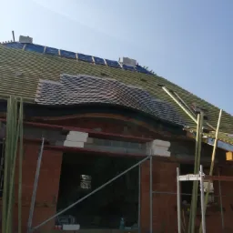 Wznoszona konstrukcja dachu z częściowo ułożoną dachówką karpiówką, widoczne elementy więźby i rusztowania przy murowanej ścianie budynku w trakcie budowy.