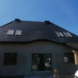 Nowy, czarny dach z dachówką bitumiczną w kształcie rybiej łuski, z oknami dachowymi, na budynku w trakcie wykończenia elewacji, z widoczną drabiną i fragmentem samochodu.