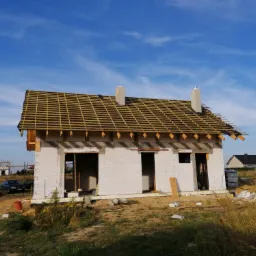 Budowa domu jednorodzinnego: ściany z białego bloczka i drewniana więźba dachowa na tle błękitnego nieba z widocznymi kominami.