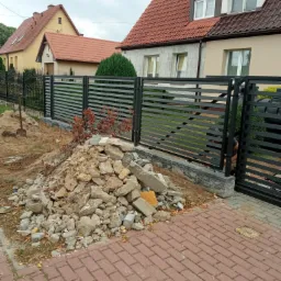 Nowoczesne, czarne ogrodzenie panelowe z poziomych listew, w trakcie montażu, widoczny stos gruzu i łopata, domy w tle.