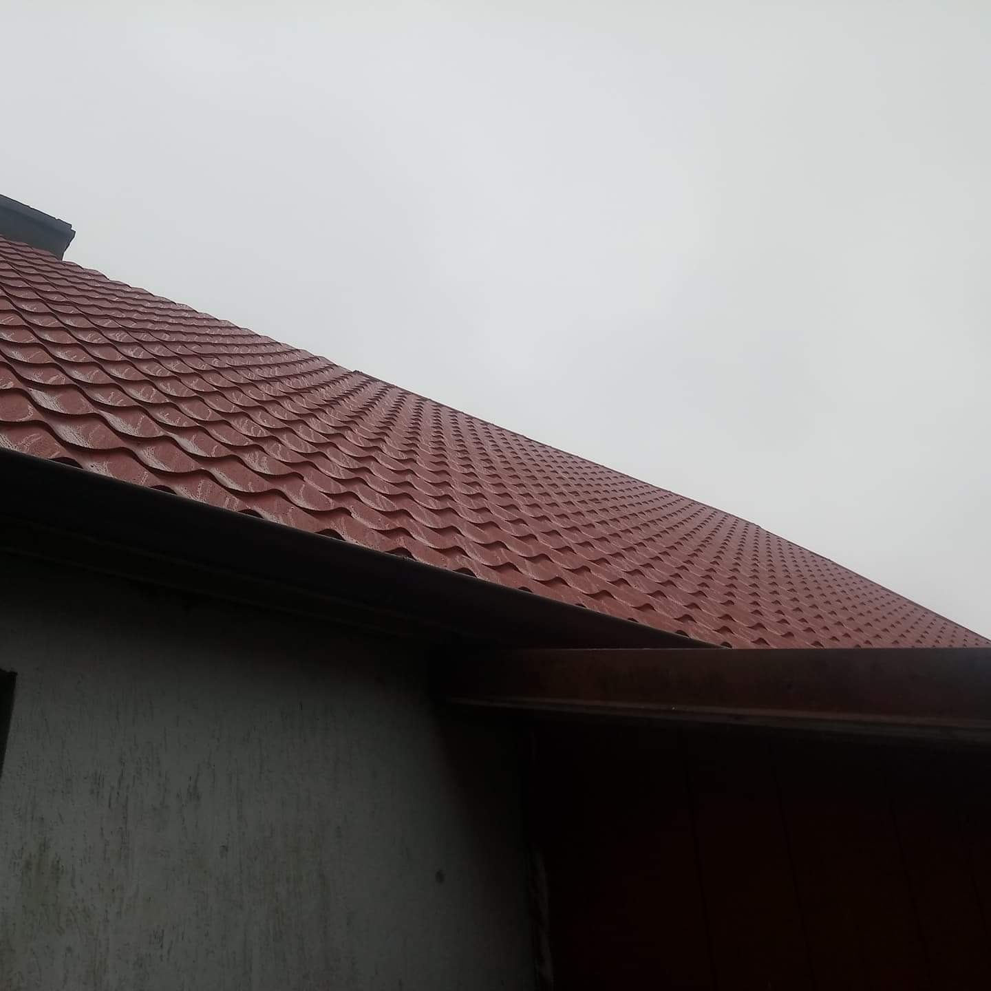 Czerwony dach z blachodachówki modułowej, widok z dołu na fragment elewacji budynku i pochmurne niebo.