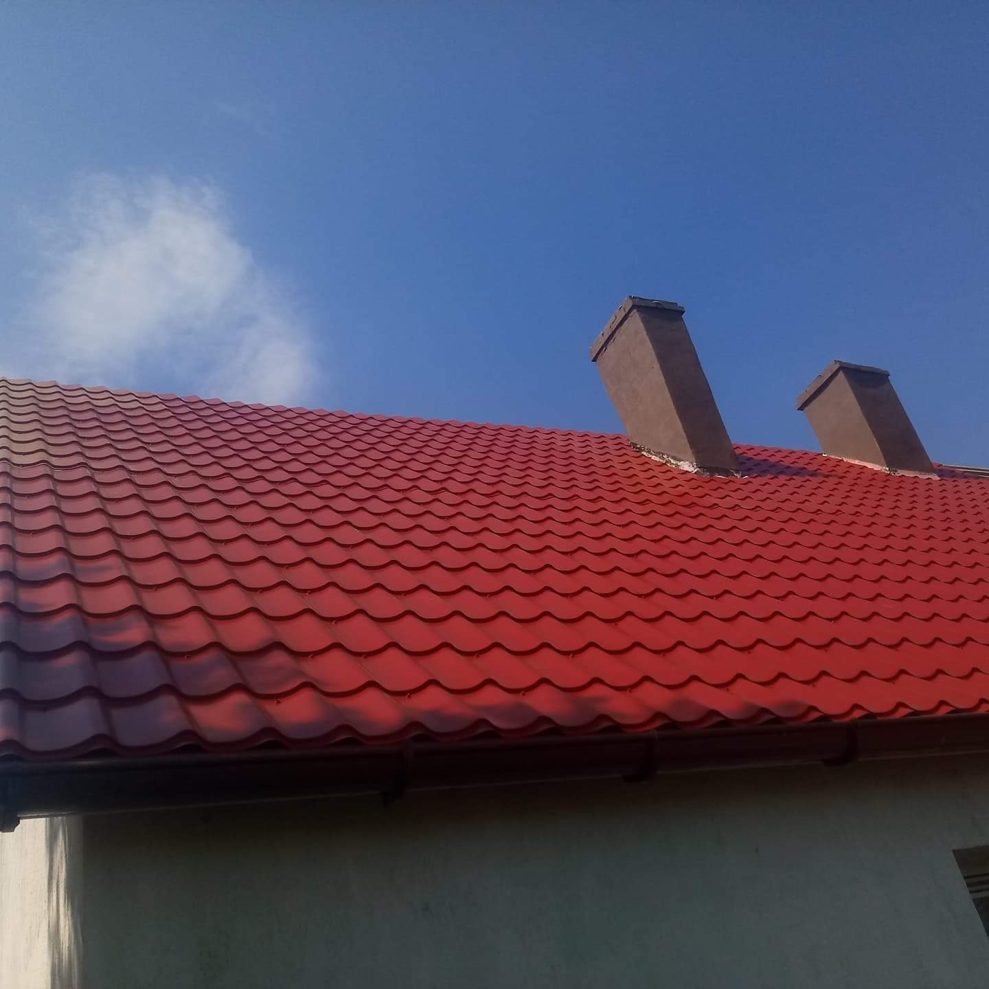 Czerwony dach z blachy trapezowej z dwoma kominami na tle błękitnego nieba z pojedynczym obłokiem.