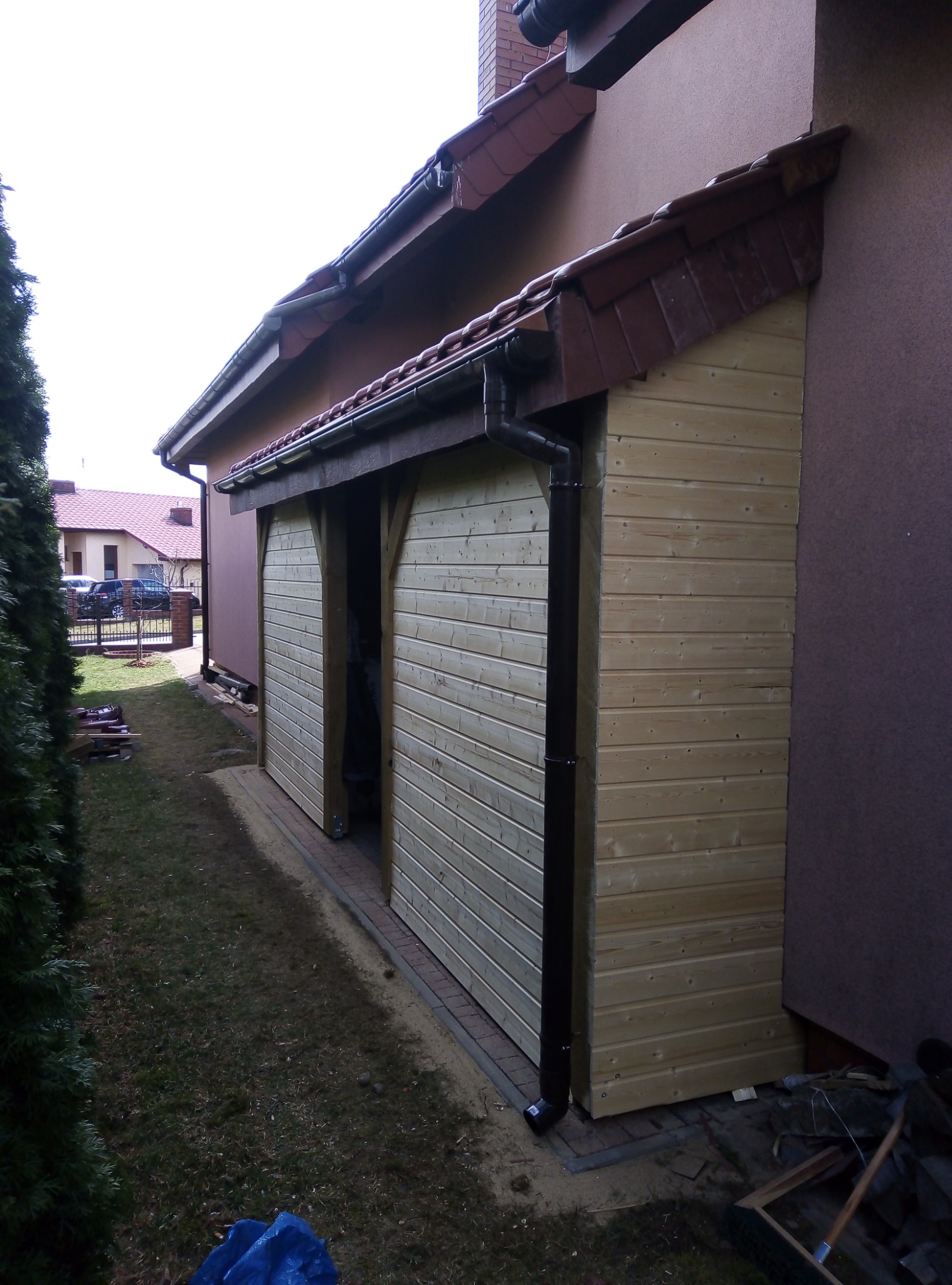 Drewniana konstrukcja z przesuwnymi drzwiami, prawdopodobnie garaż lub pomieszczenie gospodarcze, przylegająca do budynku z brązową elewacją i ciemnoczerwoną dachówką, widoczny system rynnowy