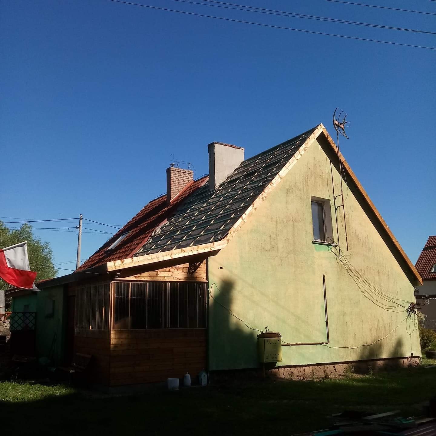 Dom w trakcie wymiany dachu, widoczne krokwie i częściowo ułożona dachówka, ocieplona elewacja w kolorze pistacjowym, dobudowany drewniany taras z polską flagą w tle.