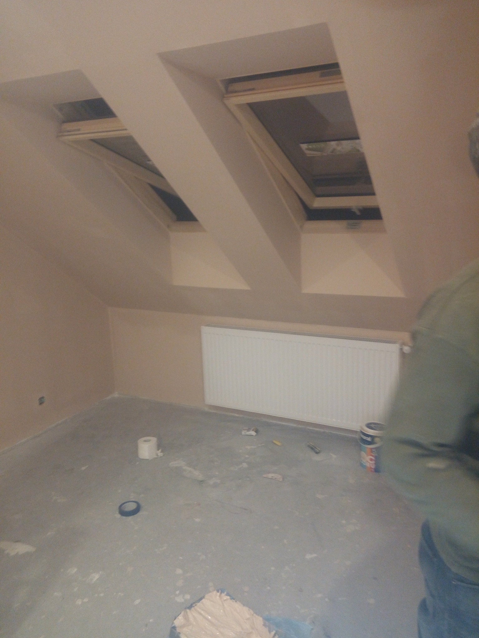 Poddasze po gładzi, widoczne dwa okna dachowe, grzejnik, wiadro z gładzią i akcesoria malarskie na podłodze, częściowo widoczna sylwetka osoby.