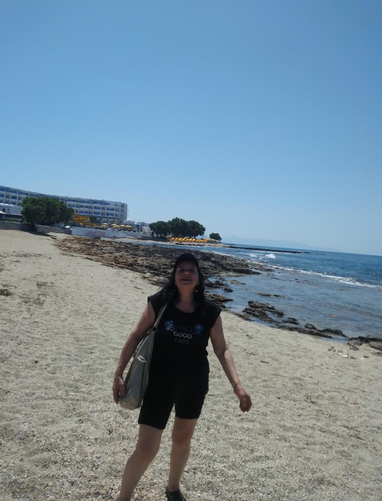 Kobieta na plaży w czarnym stroju i torbą na ramieniu, w tle błękitne morze, budynek i niebo. Słoneczny dzień.