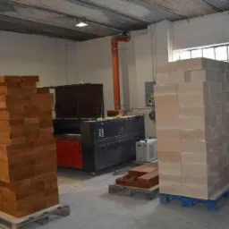 Magazyn z ułożonymi na paletach pakietami drewnianych elementów, w tle widoczna maszyna do obróbki drewna i instalacja wentylacyjna.