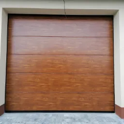 Zamknięta segmentowa brama garażowa w kolorze jasnego drewna, z widoczną strukturą słojów, zamontowana w jasnej elewacji budynku z brązowym cokołem. Po lewej stronie widoczny fragment białego krzesła.