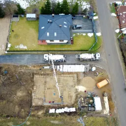 Widok z lotu ptaka na plac budowy domu jednorodzinnego, z fundamentami w trakcie wylewania betonu, dwoma betonomieszarkami i pompą do betonu, w tle gotowy dom z ogrodem.