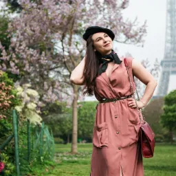 Kobieta w r&oacute;żowej sukience z kieszeniami i czarnym berecie pozuje na tle wieży Eiffla w Paryżu, z zielonym paskiem i czerwoną torebką na łańcuszku.