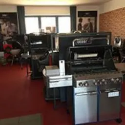 Wnętrze salonu z ekspozycją grilli gazowych i węglowych, na czerwonej podłodze, z ceglaną ścianą w tle i plakatami reklamowymi na ścianach bocznych.