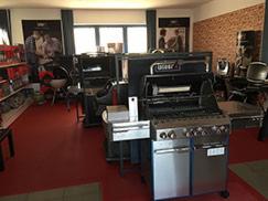 Wnętrze salonu z ekspozycją grilli gazowych i węglowych, na czerwonej podłodze, z ceglaną ścianą w tle i plakatami reklamowymi na ścianach bocznych.