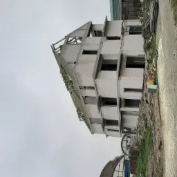 Surowy stan trzypiętrowego budynku z białych pustaków, z widoczną konstrukcją dachu, na tle pochmurnego nieba i częściowo widocznych innych budynków.