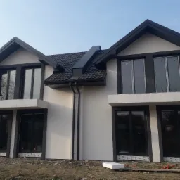 Nowoczesny dom dwukondygnacyjny z białymi ścianami i czarnymi ramami okiennymi oraz ciemną dachówką, widoczne rynny i fragment terenu przed budynkiem.