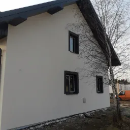 Świeżo otynkowany dom jednorodzinny z ciemnymi ramami okiennymi i elewacją w kolorze złamanej bieli, widoczny fragment brzozy na pierwszym planie i pomarańczowy samochód dostawczy w tle.