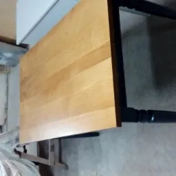 Blat drewniany z czarnymi nogami, ustawiony w warsztacie, widoczny fragment osłony z białym materiałem w tle.