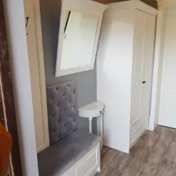 Biały przedpokój z siedziskiem tapicerowanym szarą tkaniną, lustrem w białej ramie i szafą dwudrzwiową, uzupełniony o półokrągły stolik.