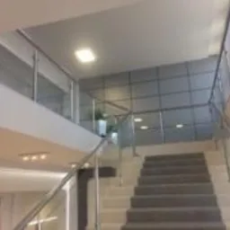 Wnętrze biura: schody z metalową balustradą, szara ściana z panelami, oświetlenie sufitowe, roślina doniczkowa na antresoli.