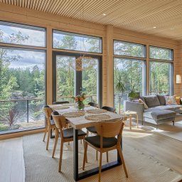 Kuusamo Polska Domy z Bali - Jasne wnętrze domu z bali z dużymi oknami wychodzącymi na las i jezioro. Widoczny salon z jadalnią, drewniane wykończenie i nowoczesny kominek.