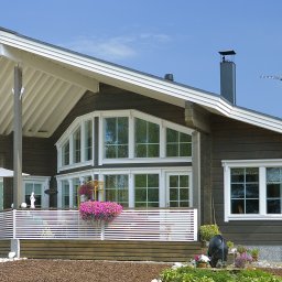 Kuusamo Polska Domy z Bali - Elegancki dom z bali w stylu skandynawskim z dużymi oknami, białymi detalami i kwiatami na balkonie. Widoczny komin i antena na dachu. Błękitne niebo w tle.