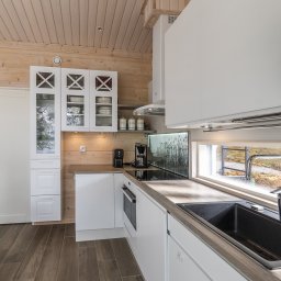 Kuusamo Polska Domy z Bali - Jasna kuchnia w domu z bali: białe szafki, drewniane ściany, okno z widokiem na zewnątrz. Nowoczesny design z elementami rustykalnymi, drewniany stół z ławką.