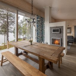 Kuusamo Polska Domy z Bali - Jasne wnętrze domu z bali z widokiem na jezioro. Duży drewniany stół i ława, kominek z telewizorem, drewniane ściany i podłoga. Nowoczesny design z naturalnymi elementami.