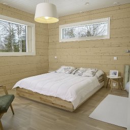 Kuusamo Polska Domy z Bali - Jasna sypialnia w domu z bali: drewniane ściany, duże okna z widokiem na drzewa, biała pościel na niskim łóżku, lampa sufitowa i fotel z zielonym obiciem.