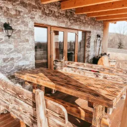 Rustykalny stół z ławkami z opalanych desek na drewnianym tarasie, z widokiem na kamienną ścianę domu i przeszklone drzwi.