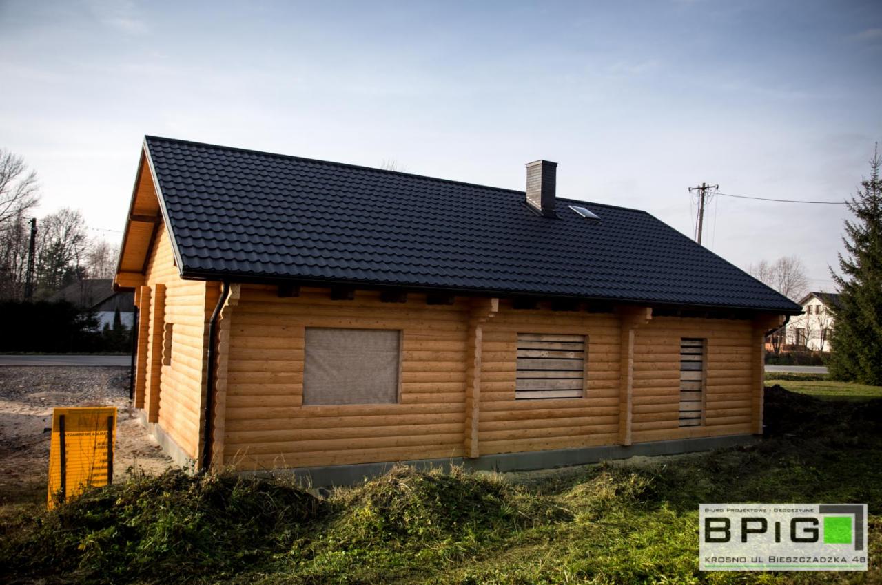 Nowo wybudowany parterowy dom z bali z ciemnym dachem z blachy, zabezpieczone okna, widoczny komin i logo firmy budowlanej w rogu.