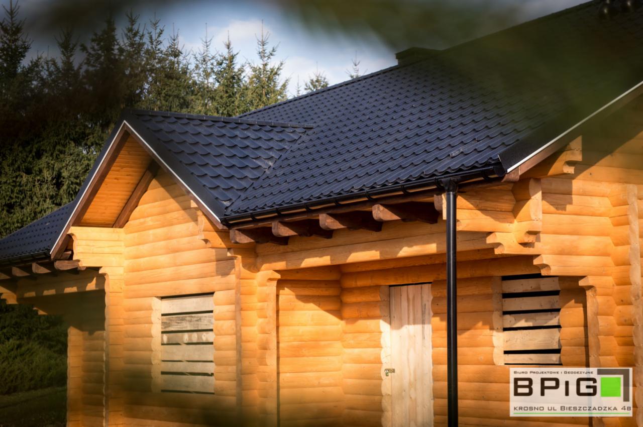 Zewnętrzny widok na drewniany dom z bali, z ciemną, metalową dachówką i deskami zasłaniającymi okna, na tle zielonych drzew.